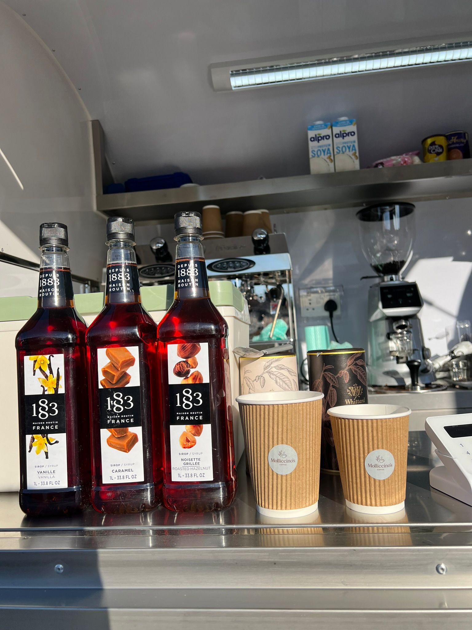 Three 1883 flavour syrups alongside Molliccino's branded cups on a sunny day